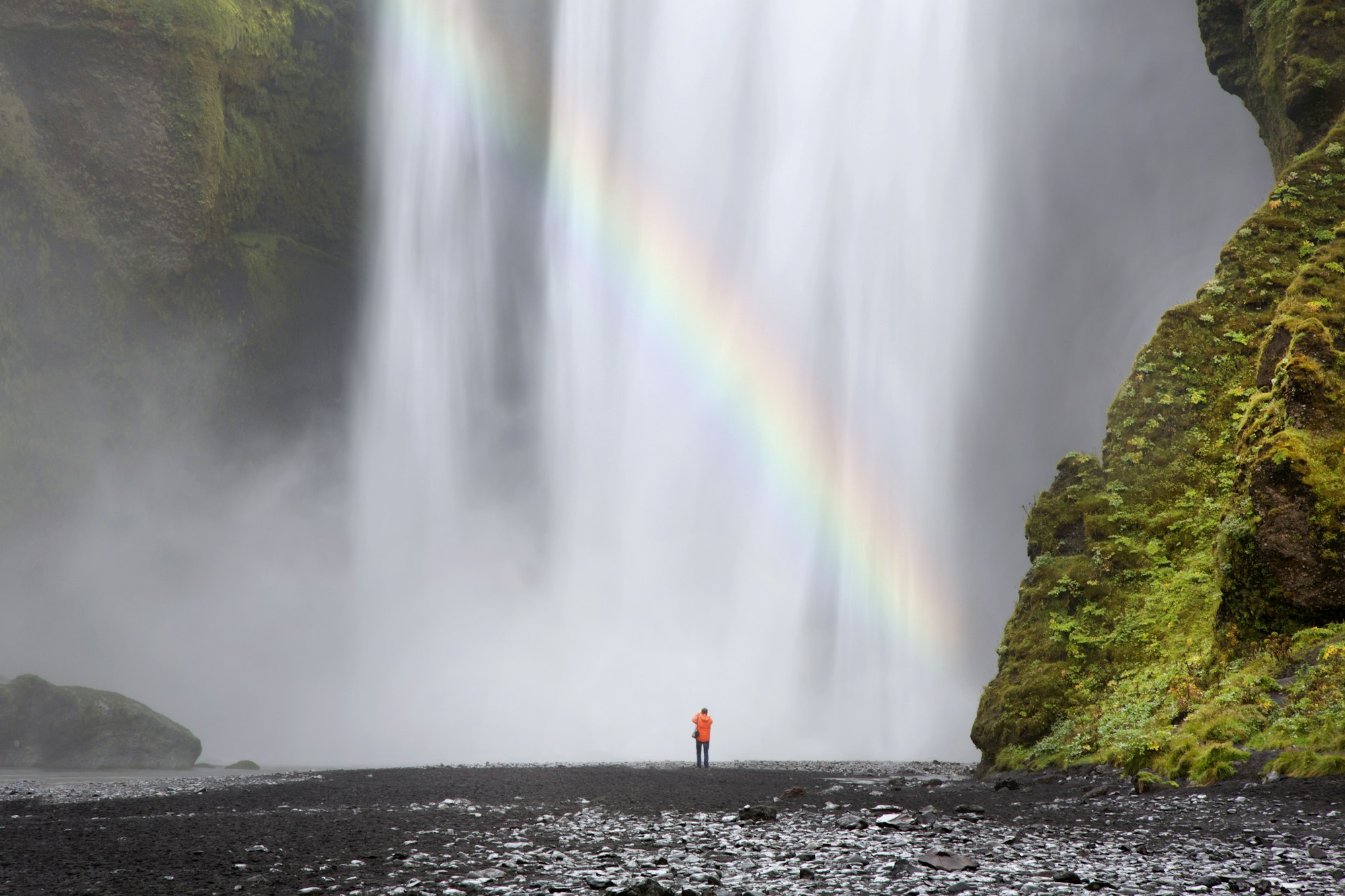 Rear view of person standing at foot of waterfall, moss-covered rocks and waterfall.